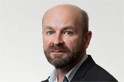 [ai] A middle-aged man with a bald head and a short beard, wearing a dark blazer and a light blue shirt, poses against a plain white background.